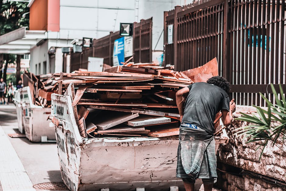 A man with curly black hair, wearing a dark t-shirt and checked shorts, is standing on a pavement next to a large, open skip filled with old wooden floorboards, broken furniture, and assorted debris, indicating a home renovation or clearing process. The skip is positioned on the street adjacent to a residential property, with a tall brown metal fence running alongside it. Behind the fence, there are signs and a building. The area is illuminated by daylight, and the scene captures the unloading or disposal stage typical of house removals or moving services. In the background, a few pedestrians are visible, but the focus remains on the man’s activity with the waste materials, which are ready for waste disposal or recycling, a common aspect of house clearance in a property transfer or relocation context. This setting reflects the logistical elements involved in packing, loading, and clearing during a home relocation managed by companies like Movers Marylebone.