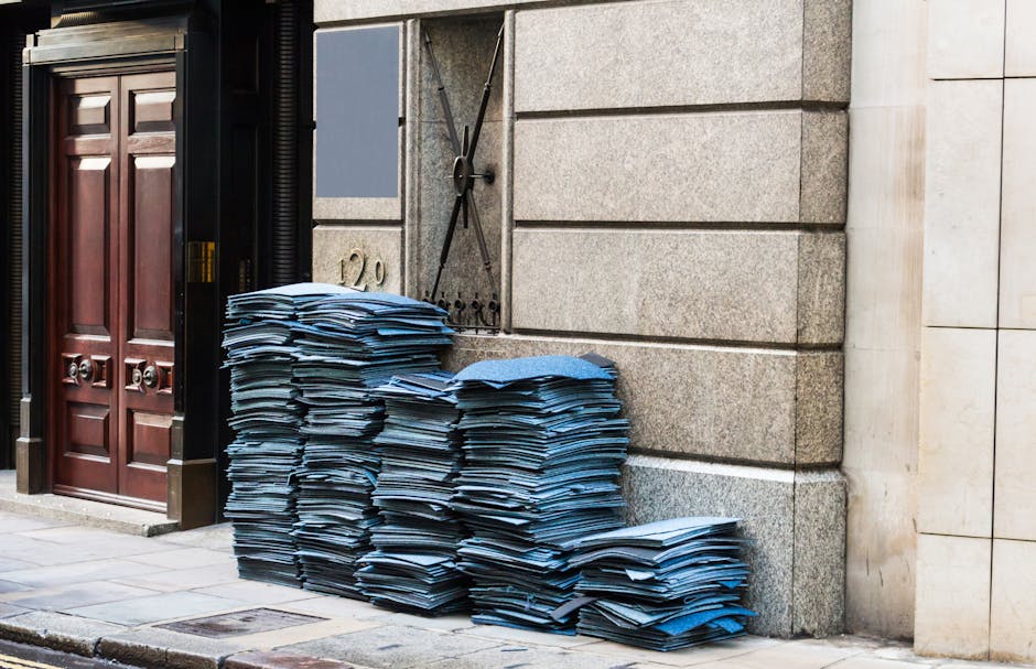 A large pile of tightly packed blue and black cardboard folders and papers stacked outside a building on a city sidewalk, positioned near a dark wooden door and a stone wall with a black metal gate. The papers appear to be old documents or files, some slightly crumpled or uneven in stacking. The scene is outdoors with natural daylight illuminating the area, and the pavement is visible in the foreground. This image illustrates household or office waste collection, which movers Marylebone can assist with during home relocations and packing and moving services, ensuring efficient clearance of bulky materials before a professional move takes place.