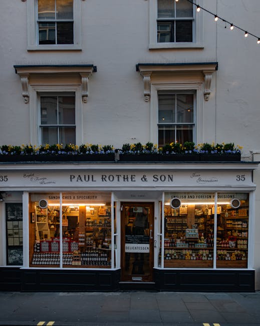 The image depicts the exterior of Paul Rothe & Son, a shop located at number 35 on a street in Marylebone, with a white facade and large display windows showcasing a variety of packaged goods such as jars, boxes, and bottles. Above the store, there is a decorative window balcony with a row of yellow and purple flowers in a planter box. The shop's sign reads 'Paul Rothe & Son' and indicates it specializes in sandwiches, delicatessen items, and foreign provisions. The entrance features a wooden door with a small sign indicating 'Delicatessen,' and the shop is illuminated with warm lighting that highlights the interior product displays. The presence of the shop's clean pavement frontage and the architectural details of the upper windows suggest an urban environment suitable for house removals and moving services, such as those offered by Movers Marylebone, who may assist with home relocation or furniture transport in the area.