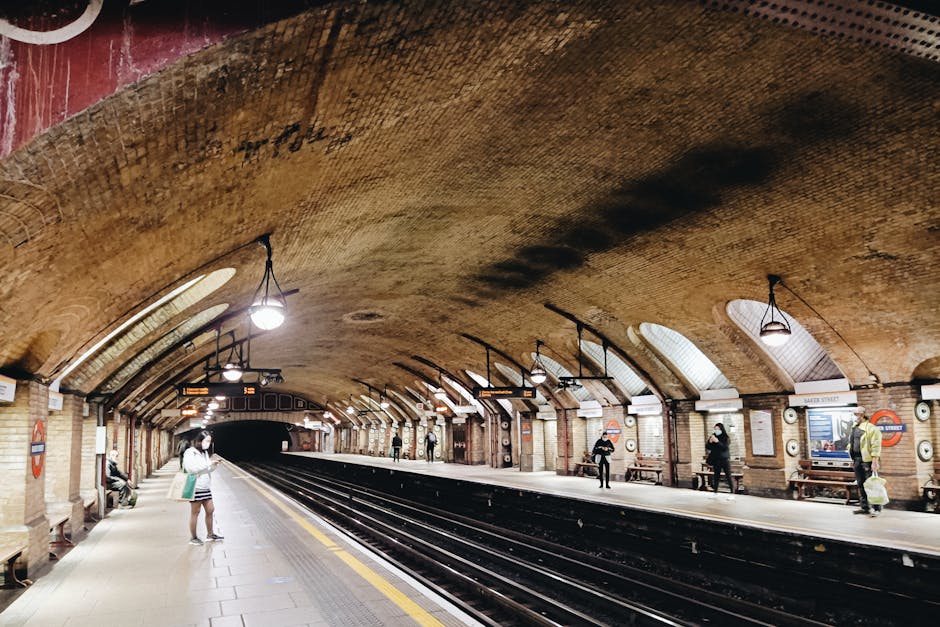 Inside Marylebone Underground Station, the brick arched ceiling is illuminated by multiple ceiling lights hanging from black fixtures, creating a well-lit environment for commuters. The platform features a yellow safety line along the edge and has a smooth tiled surface. There are several passengers waiting, some standing and others walking, with one person near the left side of the image wearing a face mask, holding a shopping bag. Bench seating is placed along the platform, with a few individuals seated. On the right, a station employee is dressed in a high-visibility jacket, standing near a luggage trolley. The station's sign displays the station name, and periodical poster boards are mounted on the wall with seating areas beneath. The overall setting is clean and organized, suggesting a routine day of passenger movement, supporting the context of moving or transporting belongings through the station as part of a house relocation or furniture transport process, complemented by the services of Movers Marylebone.