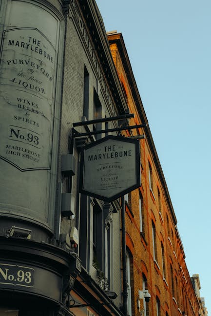 A street-level view of a historic brick building with multiple windows, featuring two black metal signs attached to the exterior: one sign is rectangular and the other is hexagonal, both displaying the name 'The Marylebone' and additional text about the address and offerings such as wine, spirits, and liquor. The signs are mounted on brackets extending from the building's façade against a clear blue sky. This exterior scene depicts the type of setting where house removals or furniture transport services by Movers Marylebone might coordinate pickups or deliveries for inner-city properties, with the signs relating to local businesses or residential buildings that could be involved in a home relocation or moving process.