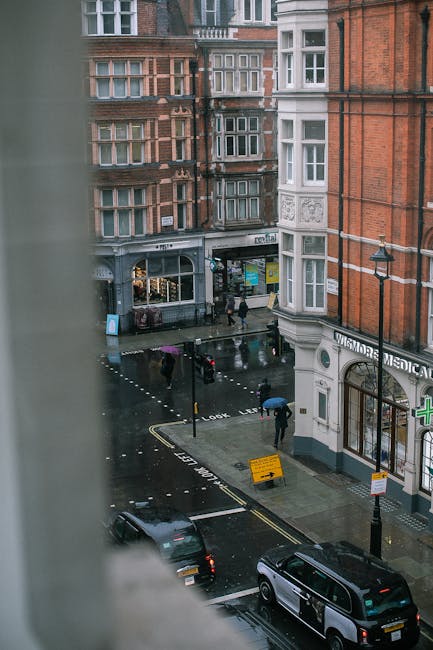 View from a window overlooking a narrow street in W1, showing the exterior of multi-storey red brick and white facade buildings with large bay windows. On the street, there are pedestrians walking under umbrellas on a rainy day, a few parked cars, and a van parked near the curb. To the right, a building houses a pharmacy with a green cross sign, and to the left, a shop with large windows and a canopy. The street has road markings and a yellow barrier indicating restricted parking or loading zones. The scene depicts a busy urban environment typical for house removals and furniture transport, with visible elements such as vehicle loading areas and pedestrians waiting, aligning with the process of packing, moving, and logistical arrangements handled by Movers Marylebone during home relocation in W1.