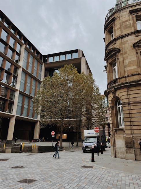 View of a narrow residential street featuring red-brick buildings with white-framed windows, in an urban setting. Two movers from Movers Marylebone are seen loading furniture and boxes into a van park