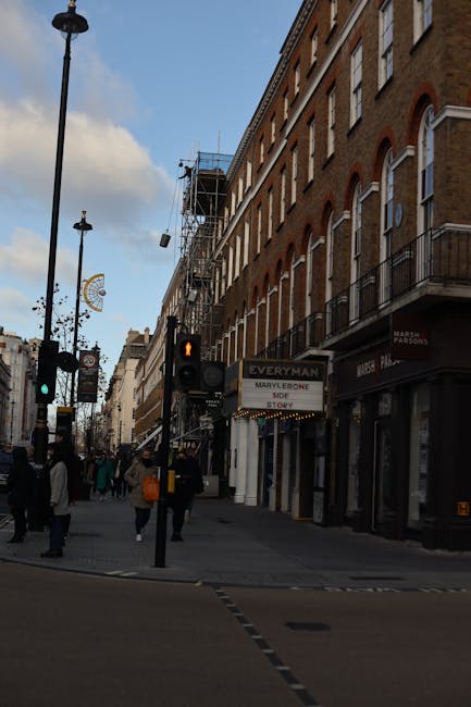 Image of a busy urban street scene in Marylebone with pedestrians crossing at a pedestrian crossing, some carrying shopping bags; a traffic signal showing amber for pedestrians; vintage-style street lamps and street signs visible along the sidewalk. A building with brick exterior walls has multiple windows and small balconies, with scaffolding on the upper floors indicating ongoing construction or renovation. The storefronts include signs for local shops, such as 'Everyman' and other retail outlets. The scene captures the movement of people and vehicles in the area, illustrating typical city activity relevant to house removals or furniture transport services, with the street environment supporting home relocation or packing and moving logistics, as provided by Movers Marylebone.