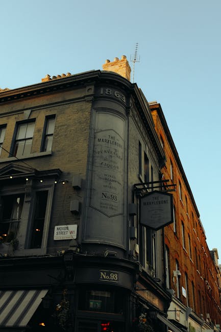 Photograph of a historic building at the corner of a street in Marylebone, London, taken during daylight with a clear sky. The building features a rounded facade with large windows and decorative architectural details, including a sign for 'The Marylebone.' The sign is mounted on a black metal bracket extending from the wall. The building is painted in beige and darker trim, with an address plate reading 'No.98' at the ground level. Nearby, a small street sign indicates 'MAYOW STREET W1.' The scene is unoccupied, and no moving or loading activities are visible in the image. The focus is on the external brickwork and signage, offering a visual context for an area where house removals and furniture transport services by Movers Marylebone might operate, especially in relation to local relocation logistics and historic street environments.