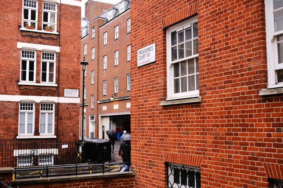 The image shows the exterior of red-brick residential buildings located on Providence Court and Balderton Street in Marylebone, with street signs visible. In the foreground, a small courtyard or loading area is partially visible, separated from the street by a low brick wall and black metal railings. Two movers from Movers Marylebone are engaged in a home relocation process; they are carrying a large piece of furniture wrapped in protective blankets and plastic wrap, likely for transport to or from a van parked nearby. The area includes a black waste disposal bin, a trolley, and moving straps, indicating an active loading or unloading scene. The setting is outdoors during daytime, with natural light illuminating the scene, highlighting the bricks of the buildings and the various objects involved in the furniture transport and packing process, which are typical aspects of professional removals and house moving services in Marylebone.